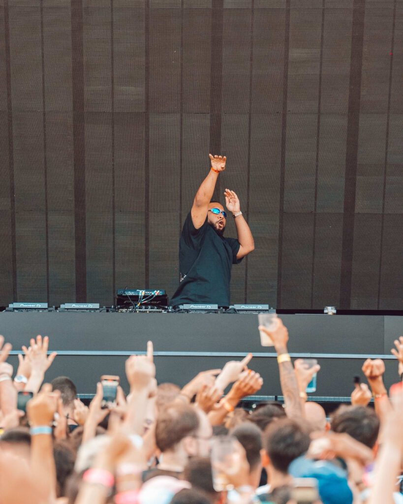 DJ performing on stage at an outdoor music festival, surrounded by a large crowd with hands raised in excitement