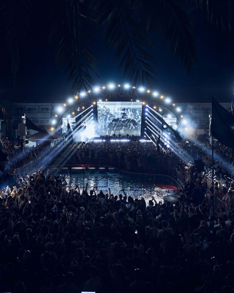 A massive crowd enjoys a night concert at an outdoor venue with a large illuminated stage and a screen displaying performers