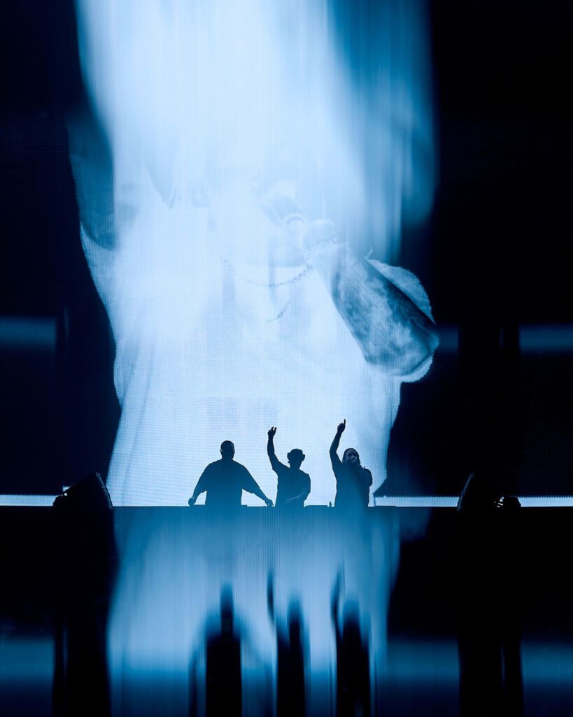 Three people stand at a DJ booth with hands raised during a live music performance, with a large projection of an arm and microphone in the background